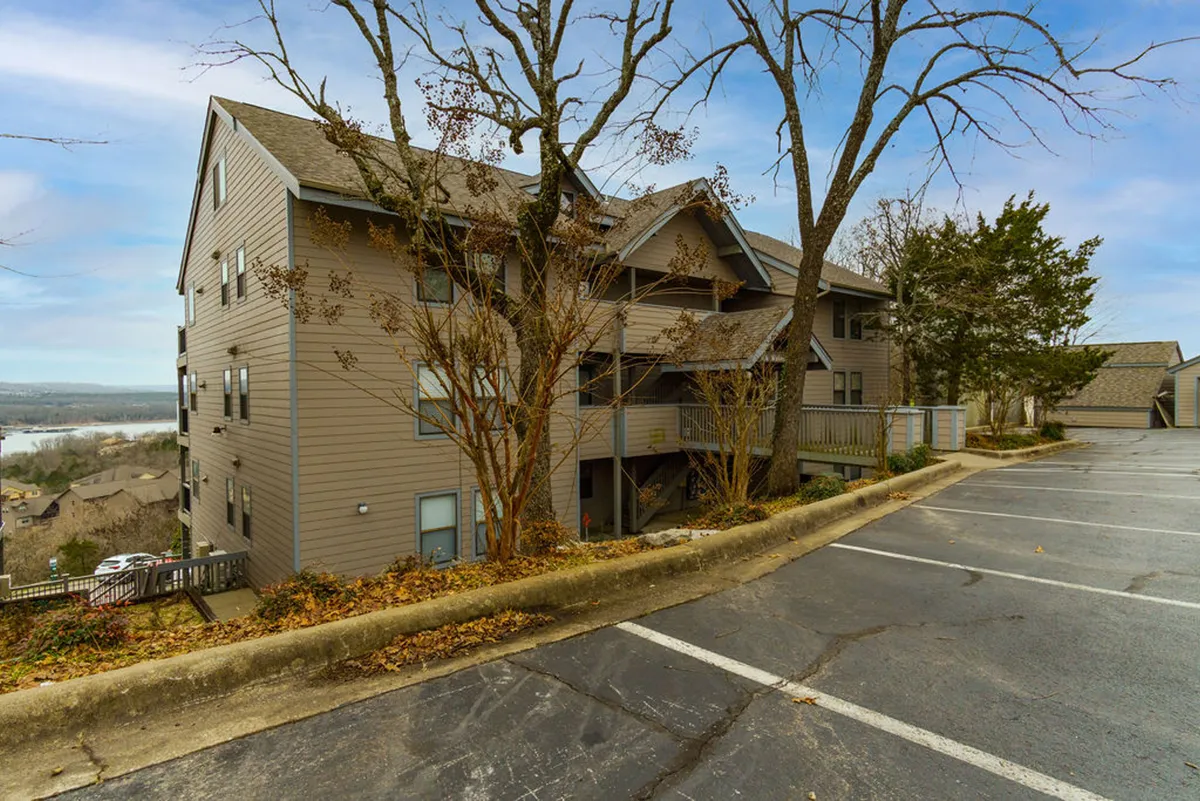A serene exterior scene showcasing the condo building's three stories, mature trees, well-kept grounds, and a glimpse of the water beyond. The setting feels peaceful and community-oriented, ideal for walks and enjoying nature.