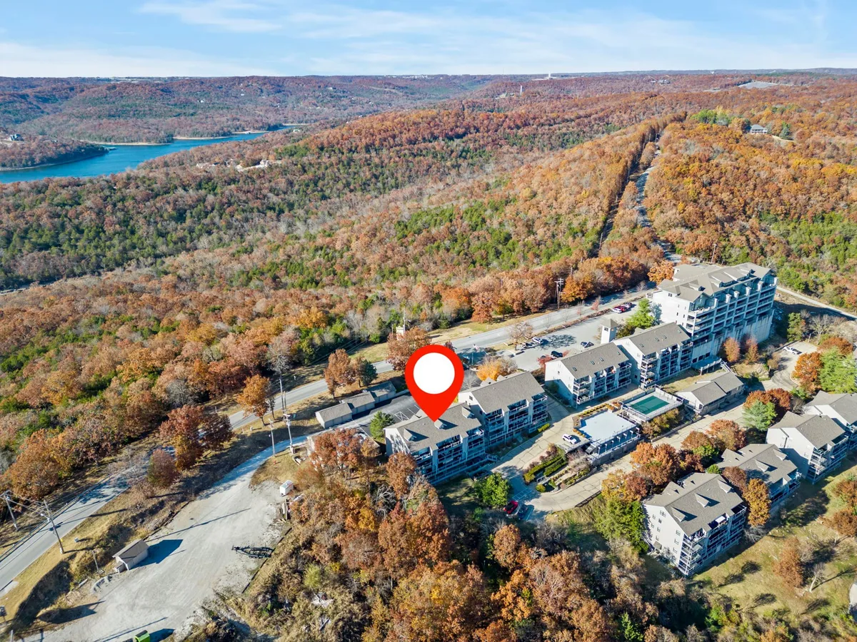 A wide view of the apartment buildings set against lush greenery, with a swimming pool visible in the courtyard and a backdrop of surrounding trees and water. The scene includes exterior communal spaces and shows the peaceful, scenic environment guests would enjoy.
