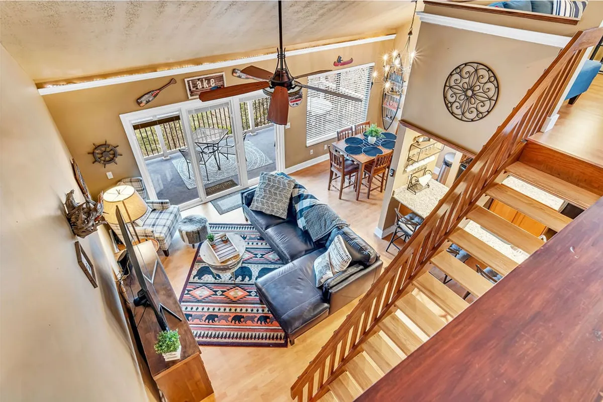The image showcases a spacious, sunlit open-concept living and dining area with cozy furnishings, a stylish area rug, and a large sliding glass door that opens to a balcony offering breathtaking lake views. The space is inviting, perfect for gathering, relaxing, and enjoying meals together while soaking in the scenery.