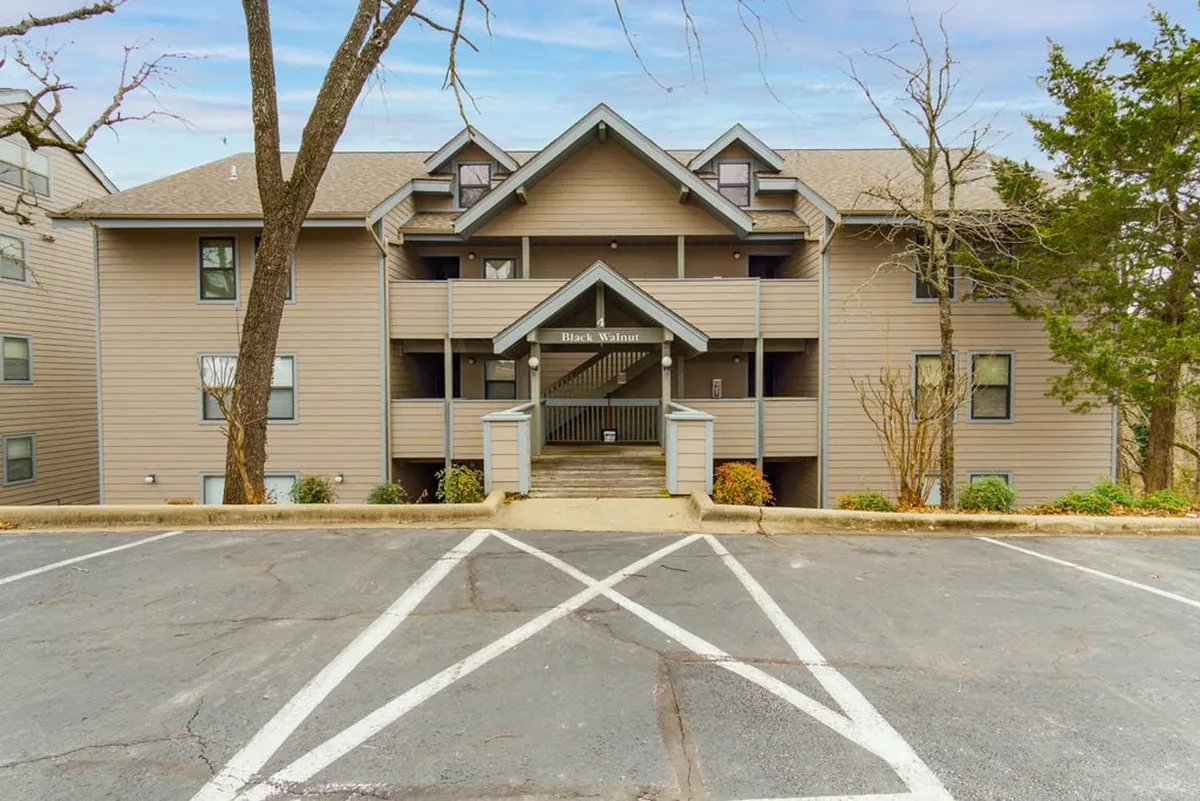 The exterior of the condo complex, showcasing the inviting front entrance, multiple balconies, landscaped grounds, and a mature tree in the foreground, all bathed in natural light.