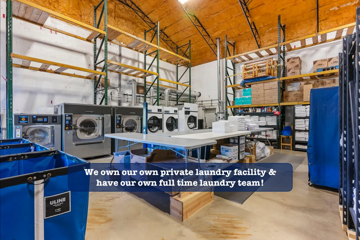 This image presents a professional, organized laundry area featuring industrial washing machines, laundry carts, and storage shelves. The environment appears clean, efficient, and well-maintained, suggesting attention to detail and high standards for guest cleanliness.
