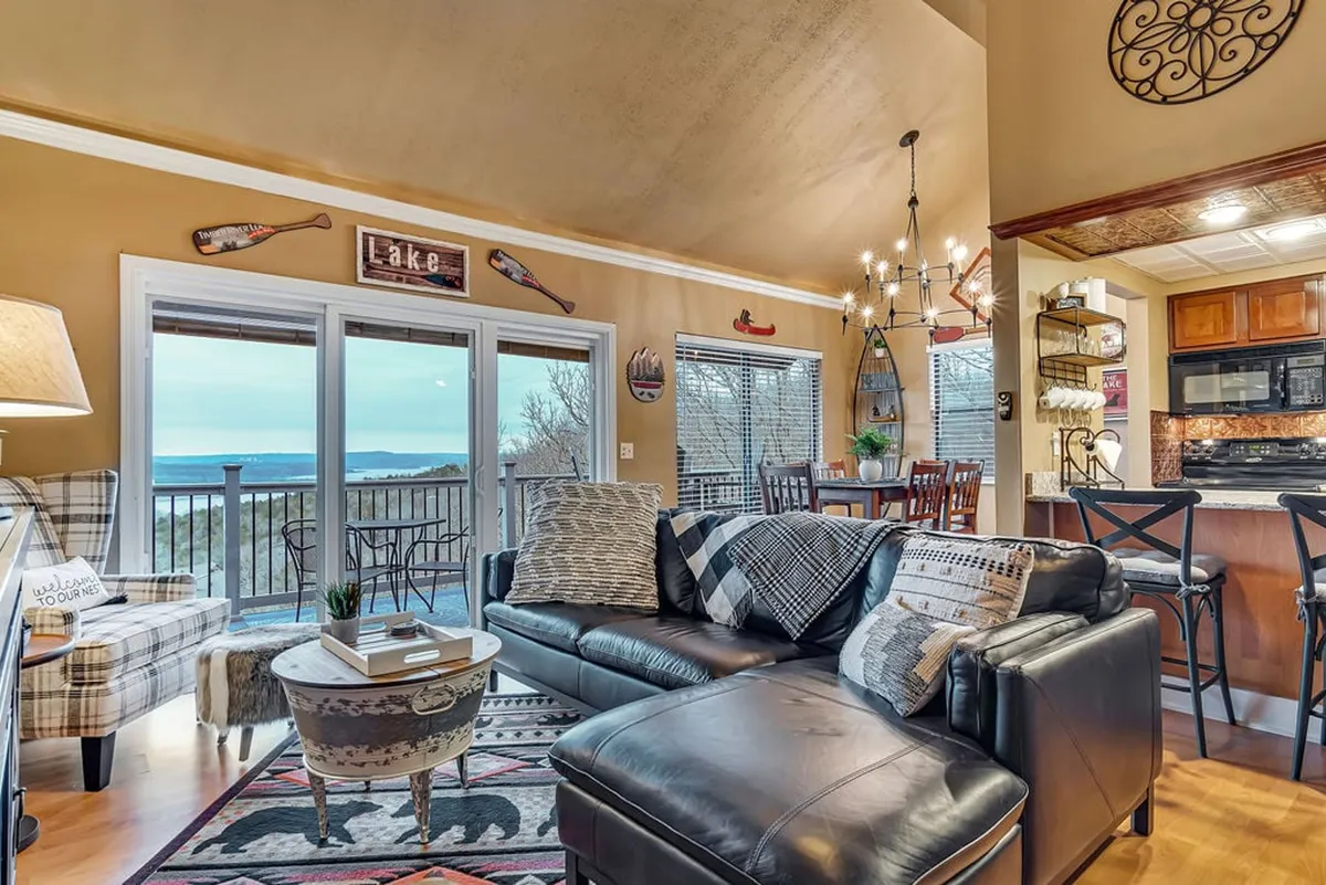 A spacious living and dining area with large windows that offer a sweeping view of Table Rock Lake. The room features a comfortable black leather sofa, a wooden dining table, and a decorative chandelier, all awash in inviting natural light. The setting suggests a seamless flow between relaxation and dining, and the scenery outside is an ever-present backdrop.